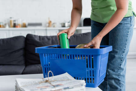 Cropped view of woman putting cans in basket near blurred newspapers at homeの写真素材