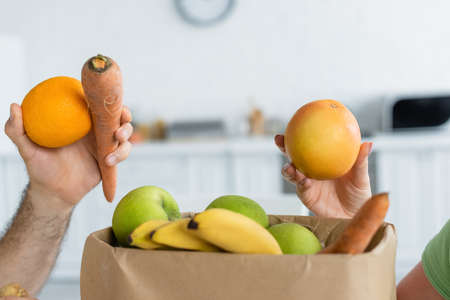Cropped view of couple holding fruits near paper bag in kitchenの写真素材
