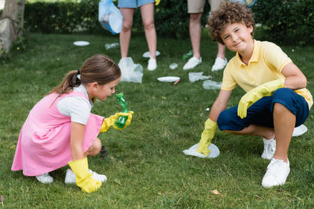 Smiling boy in rubber gloves looking at camera near garbage on lawn and family outdoorsの写真素材