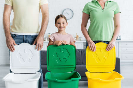 Smiling girl looking at camera near parents and trash bins with recycle signの写真素材