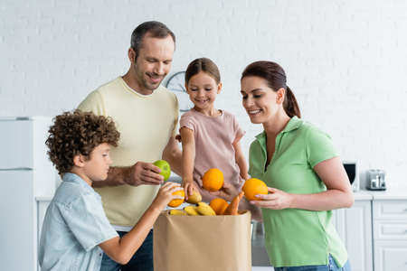 Cheerful family with kids holding fruits near paper bag in kitchenの写真素材