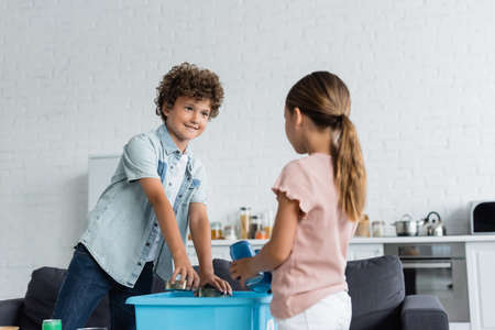 Smiling boy putting tin cans in box near blurred sister at homeの写真素材