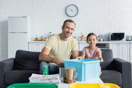 Cheerful father and daughter holding box with recycle sign near trash on table in kitchenの写真素材