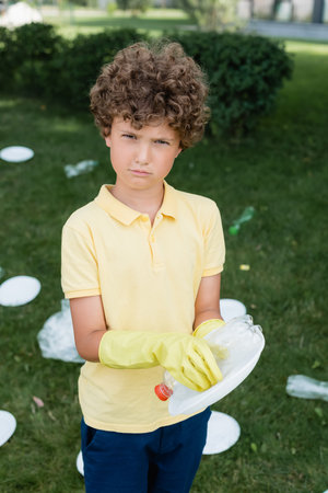 Boy in rubber gloves holding trash and looking at camera outdoorsの写真素材