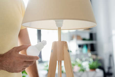 Cropped view of man holding energy saving light bulb near floor lamp at homeの写真素材