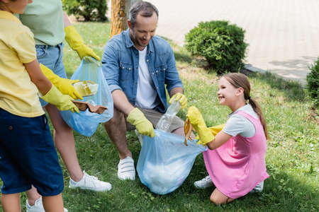 Smiling child in rubber gloves collecting trash with family on lawn
