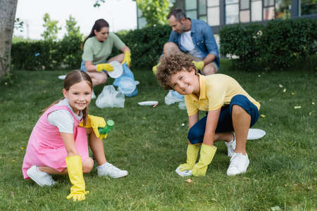 Smiling kids in rubber gloves holding trash near blurred mother and dad on lawnの写真素材