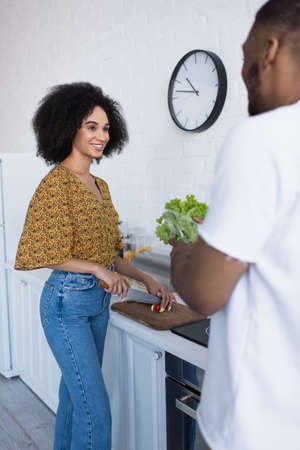 Smiling african american woman cutting cherry tomato near blurred boyfriend in kitchen