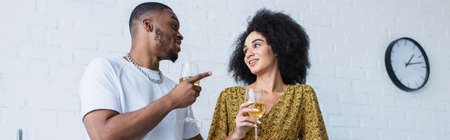 Young african american man pointing with finger while talking to girlfriend with wine in kitchen,...