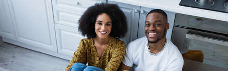 High angle view of smiling african american couple looking at camera on floor in kitchen, banner