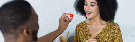 Cheerful african american woman near blurred boyfriend with cherry tomato in kitchen, bannerの写真素材