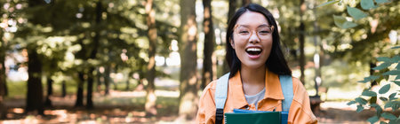 excited asian woman with notebooks laughing at camera in park, bannerの写真素材