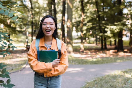 cheerful asian student laughing at camera while standing in park with copy booksの写真素材