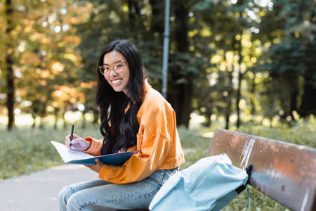 happy asian woman writing in notebook and smiling at camera on bench in parkの写真素材