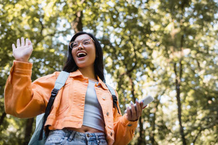 low angle view of excited asian student with backpack and smartphone waving hand in parkの写真素材