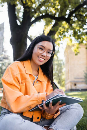 young asian woman smiling at camera while writing in notebook in parkの写真素材