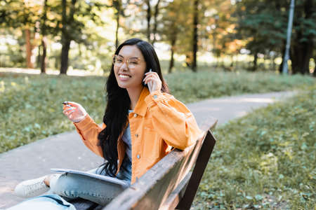 cheerful asian student with pen and notebook talking on mobile phone on bench in park