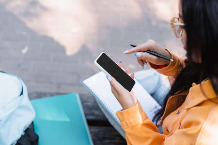 partial view of asian student using mobile phone with blank screen while sitting outdoors
