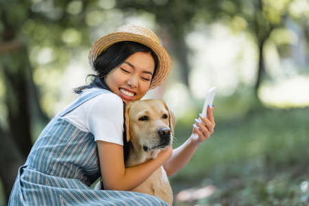happy asian woman with closed eyes hugging labrador while taking selfie in parkの写真素材