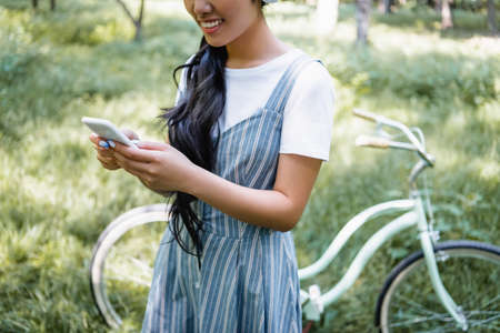 cropped view of smiling woman chatting on smartphone near blurred bike in parkの写真素材
