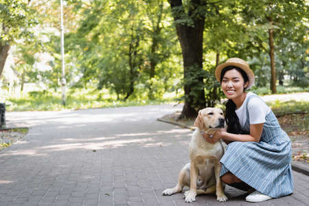 cheerful asian woman in straw hat looking at camera while hugging labrador in park