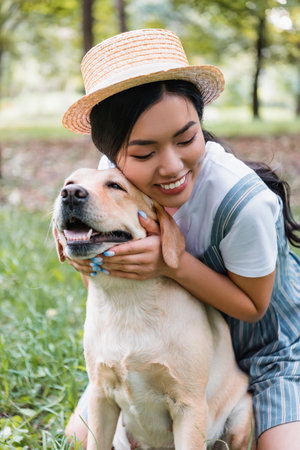 young and happy asian woman cuddling yellow labrador in parkの写真素材
