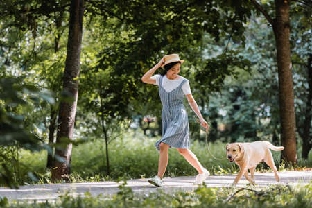 young asian woman in sundress and straw hat running with dog in parkの写真素材