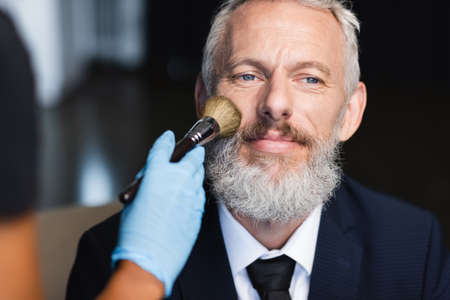 blurred african american makeup artist in latex glove applying face powder on pleased businessman in suitの写真素材