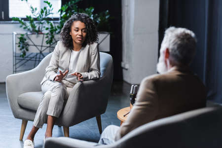 curly african american journalist gesturing while talking with blurred businessman sitting in armchair during interviewの写真素材