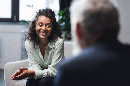 joyful african american journalist sitting in armchair and smiling near blurred guestの写真素材