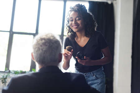 smiling african american makeup artist holding cosmetic brush near blurred businessman in interview studioの写真素材