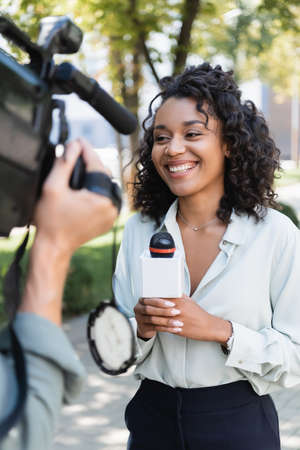 smiling african american journalist with microphone doing reportage near blurred cameraman with video cameraの写真素材