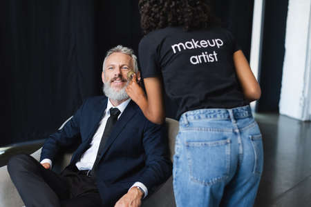 curly african american makeup artist in t-shirt with lettering applying face powder on happy businessman in interview studioの写真素材