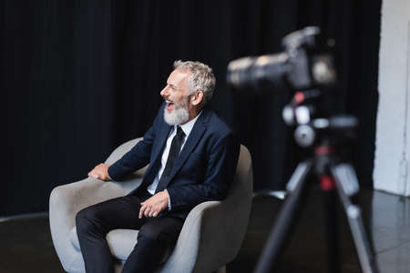 cheerful businessman in suit laughing while sitting in armchair during interview near blurred digital camera on tripodの写真素材