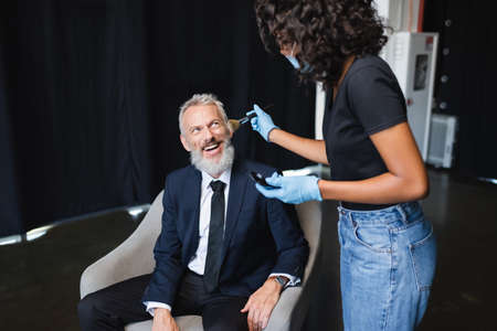 african american makeup artist in medical mask and latex gloves applying face powder on cheerful...