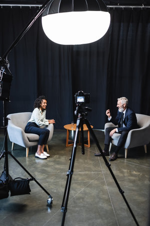 happy african american journalist sitting in armchair and talking with guest in studioの写真素材