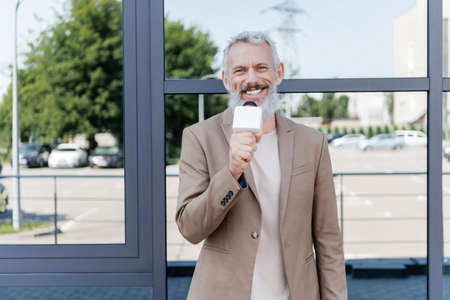 cheerful reporter in blazer holding microphone and making reportage near building