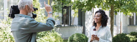 cameraman pointing with finger near happy african american reporter with microphone, banner