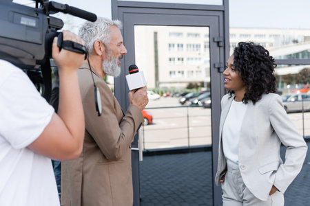 reporter with microphone interviewing happy african american businesswoman near blurred cameraman with video cameraの写真素材