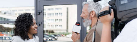 reporter with microphone interviewing happy african american businesswoman near blurred cameraman...
