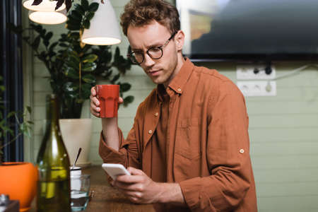 young man in glasses holding cup and texting on smartphone in cafe