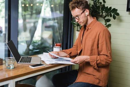 freelancer in earphones looking at graphs near gadgets and drinks on table in cafeの写真素材