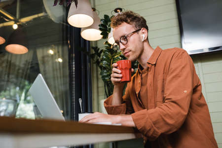 young freelancer in glasses and wireless earphones holding cup and using laptop in cafe