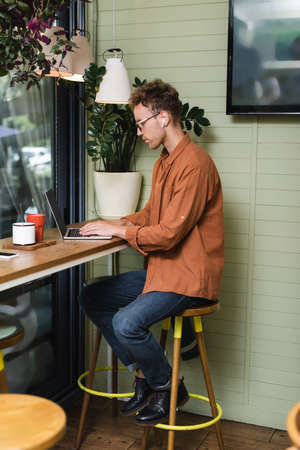 side view of young man in glasses typing on laptop in cafe