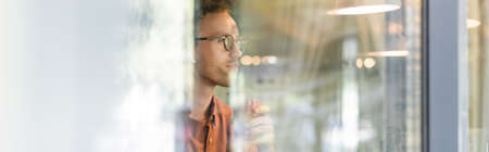 young man in glasses and wireless earphones holding cup of coffee behind blurred window of cafe, bannerの写真素材