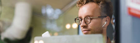 young man in glasses and earphones near laptop behind blurred window of cafe, bannerの写真素材
