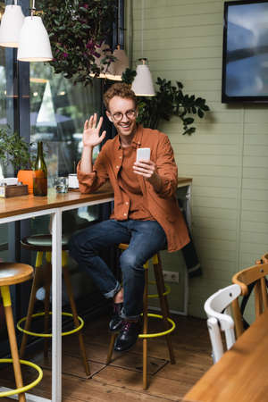 happy young man in glasses and wireless earphones waving hand during video call in cafe