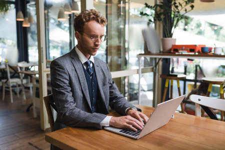 curly businessman in suit and glasses typing on laptop in cafe
