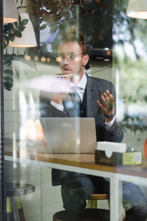 curly businessman in glasses recording voice message near laptop behind blurred window of cafe