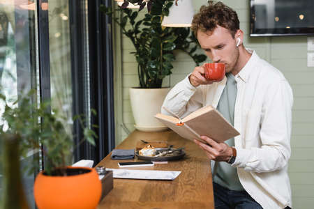 young curly man in wireless earphones reading book while drinking coffee in blurred cafe